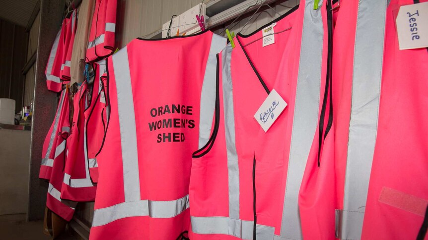 Bright pink high visibility vests hanging in a row printed with the words Orange Women's Shed, some with name tags on them