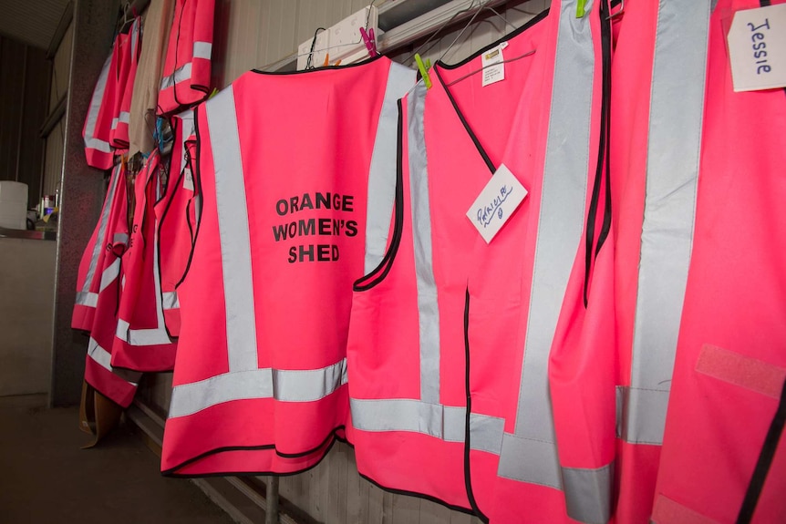 Bright pink high visibility vests hanging in a row printed with the words Orange Women's Shed, some with name tags on them