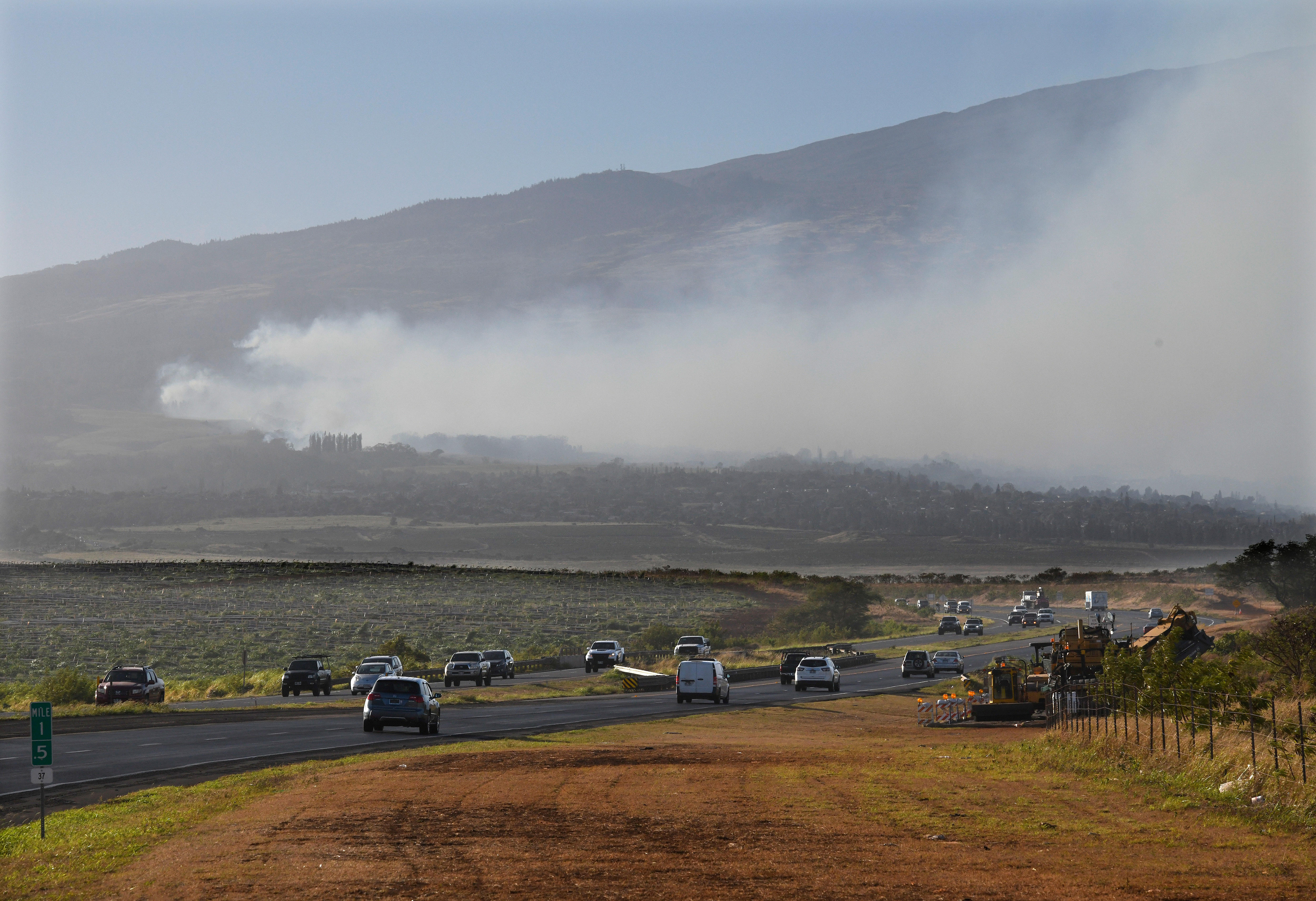 A wide shot of smoke blowing across a mountain as cars drive along a winding road in the forefront.