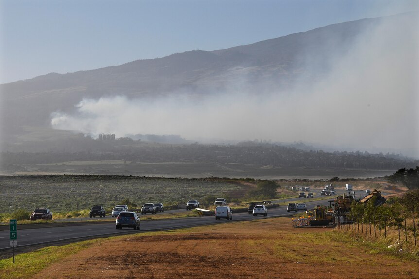 A wide shot of smoke blowing across a mountain as cars drive along a winding road in the forefront.