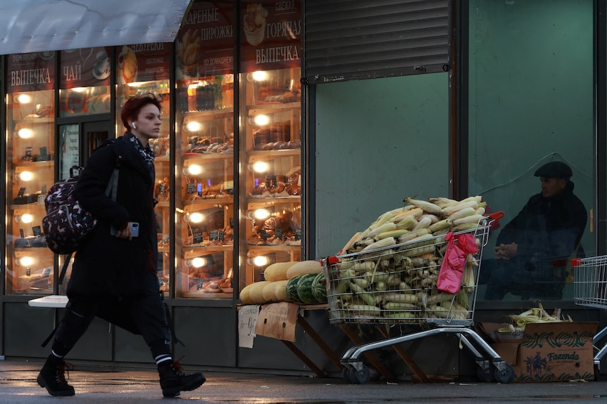 A woman in a black coat walks past a warmly-lit shop and a street vendor with a trolley full of bananas.