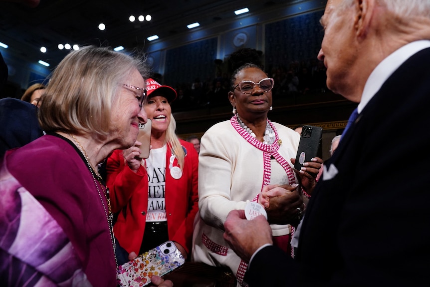 Marjorie Taylor Greene dressed in a red shirt and wearing a MAGA hate attempts to speak to Joe Biden.