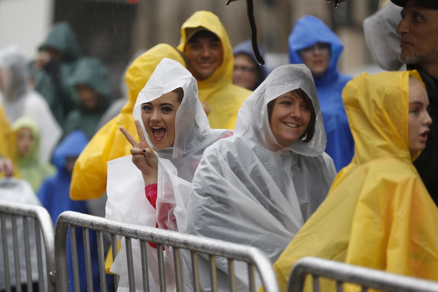 People smile towards the camera while lining up behind crowd control barriers. They're wearing rain ponchos