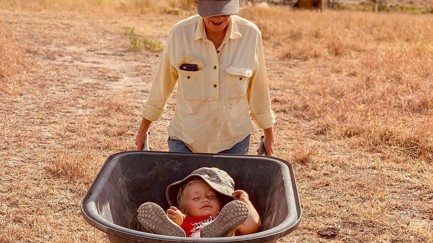 A female pushes a child who's asleep in a wheelbarrow.