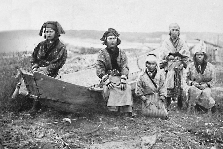 A black and white portrait shows five women in traditional dress sitting on a wooden fishing boat on the grass