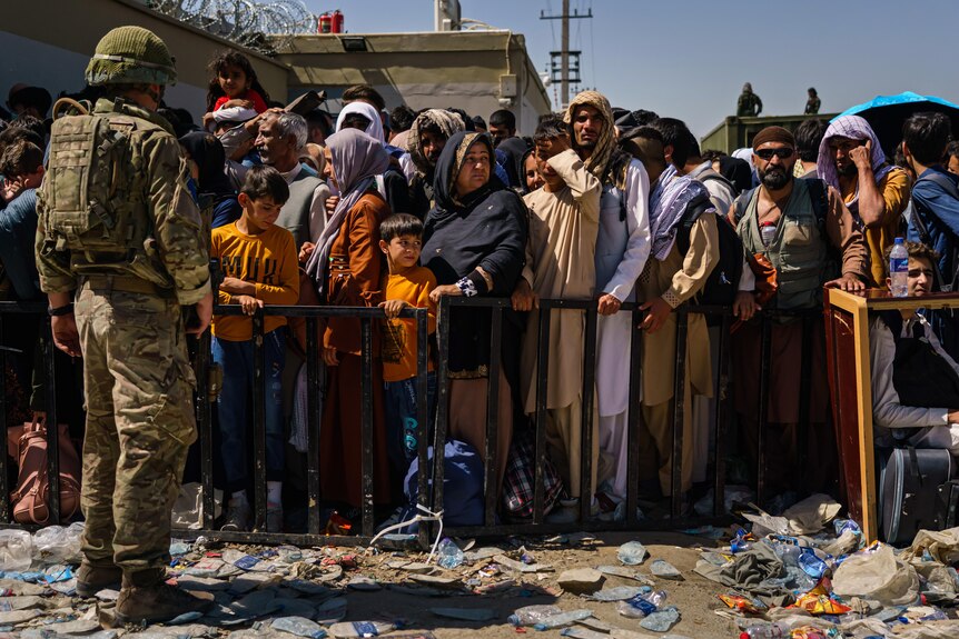 A soldier stands looking at dozens of men, women, and children standing behind a barrier. The people look concerned and tired.