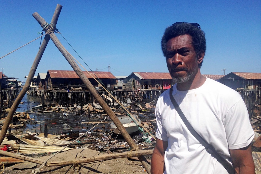 Ako Nou, a resident of Hanuabada Village. He is standing near the shore. The water is dark and littered with debris.