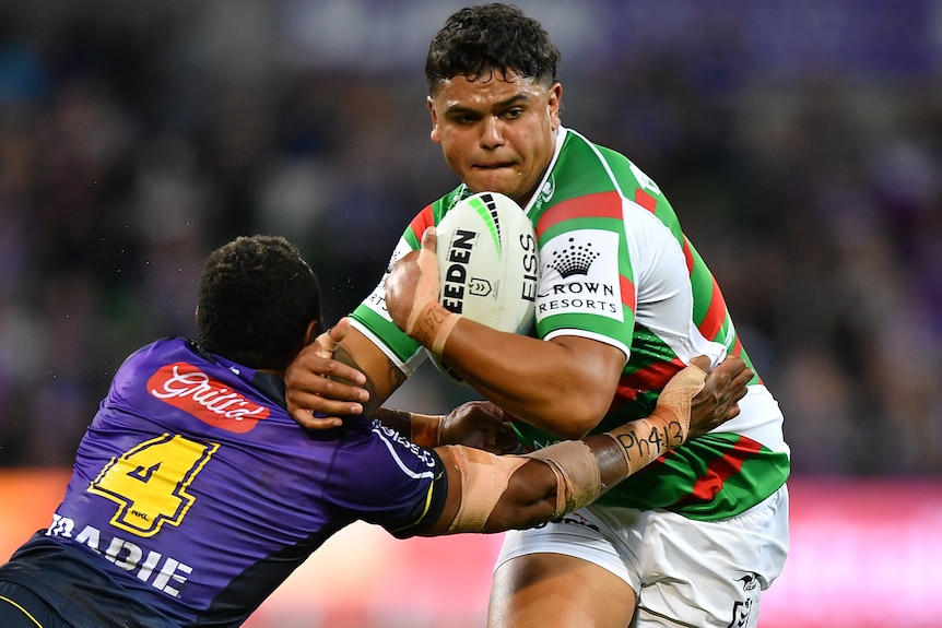 A South Sydney NRL player carries the ball as he is tackled by a Melbourne Storm opponent.