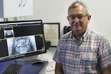 A man wearing a checked shirt sits smiling at the camera, to his left a computer screen with an x-ray of a mouth