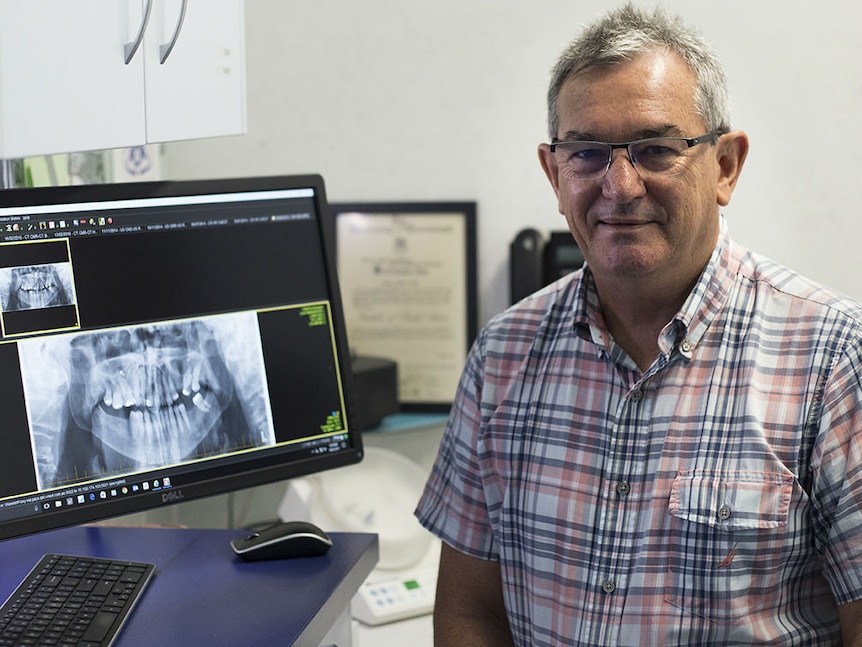 A man wearing a checked shirt sits smiling at the camera, to his left a computer screen with an x-ray of a mouth