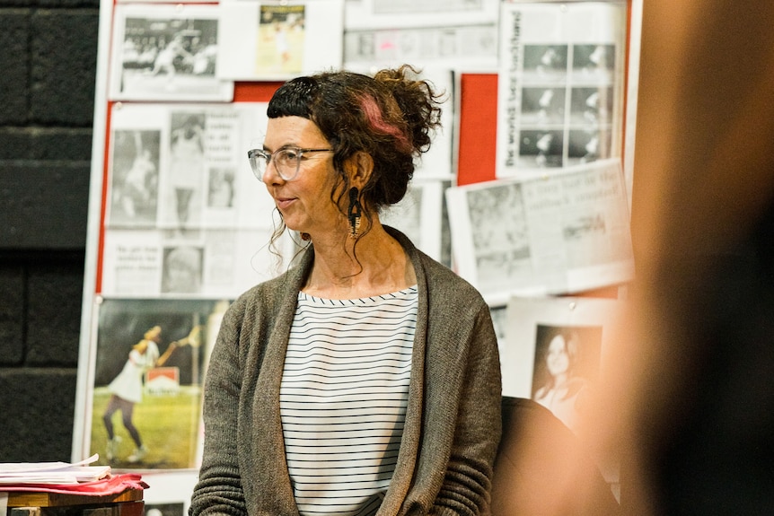 A woman wearing glasses sits with her curly hair pulled back in a bun. Behind her is a board filled with newspaper cuttings.