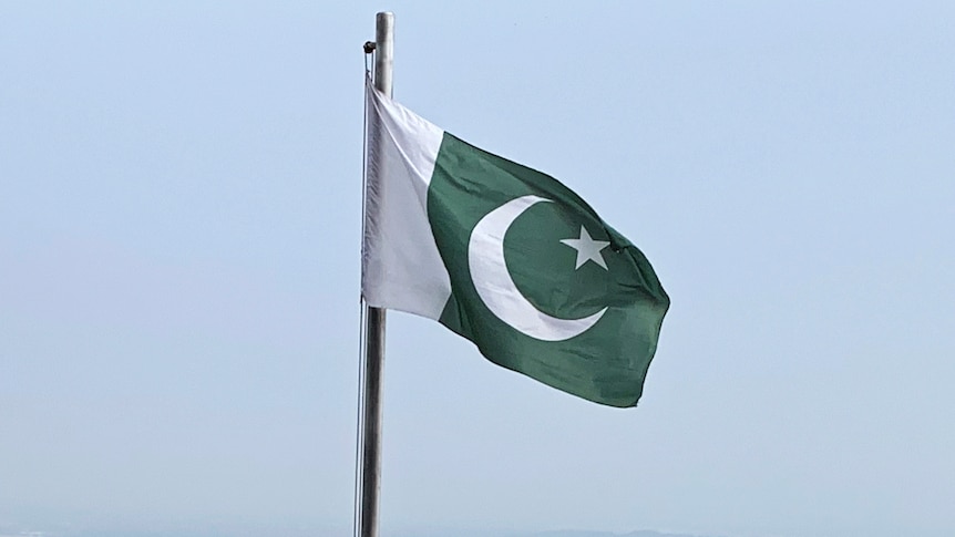 A photo of a Pakistani flag on a pole. It is green and white and has a crescent moon and star on it.