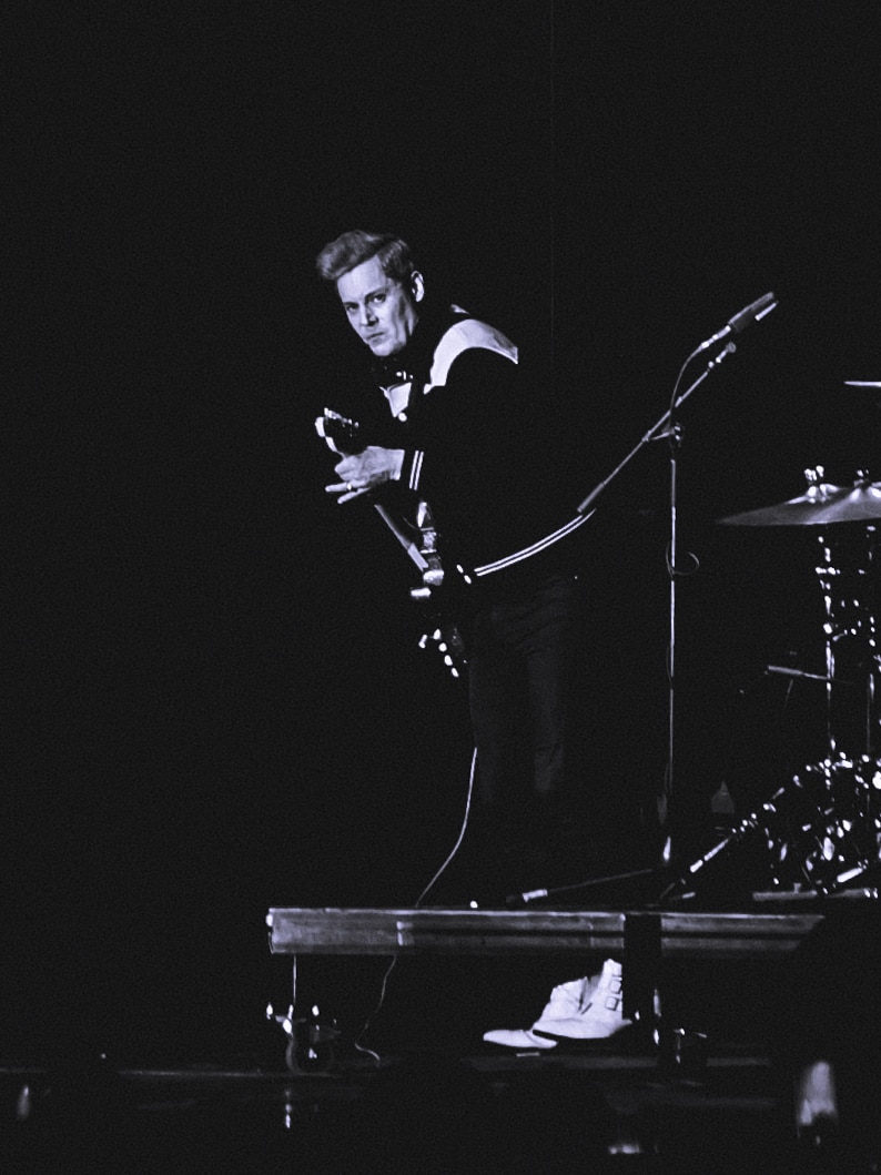 black and white photo of man in jacket playing a guitar on stage