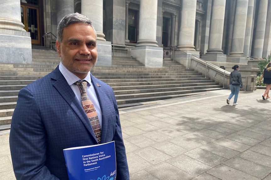 An aboriginal man in a blue suits smiles in front of a grey parliament building