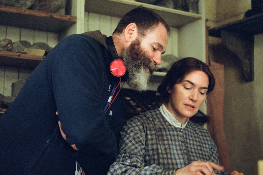 Man with bushy grey-brown beard leaning over Kate Winslet from behind as she sits at desk, in costume.