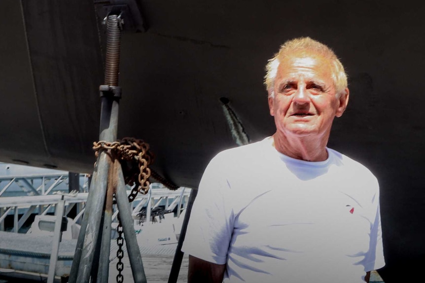 A man in a white t-shirt stands in front of a yellow and orange yacht, squinting at the sun.