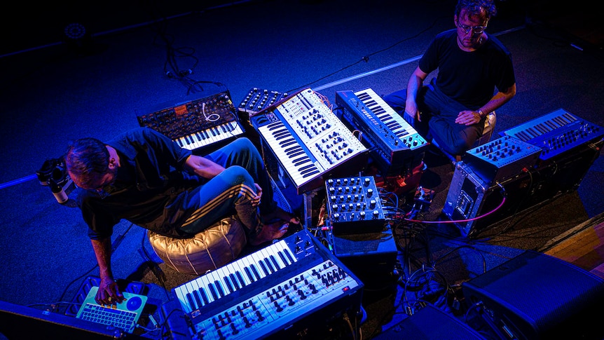 we look down on two men in a nest of analogue synthesisers and keyboards, washed in blue stage light