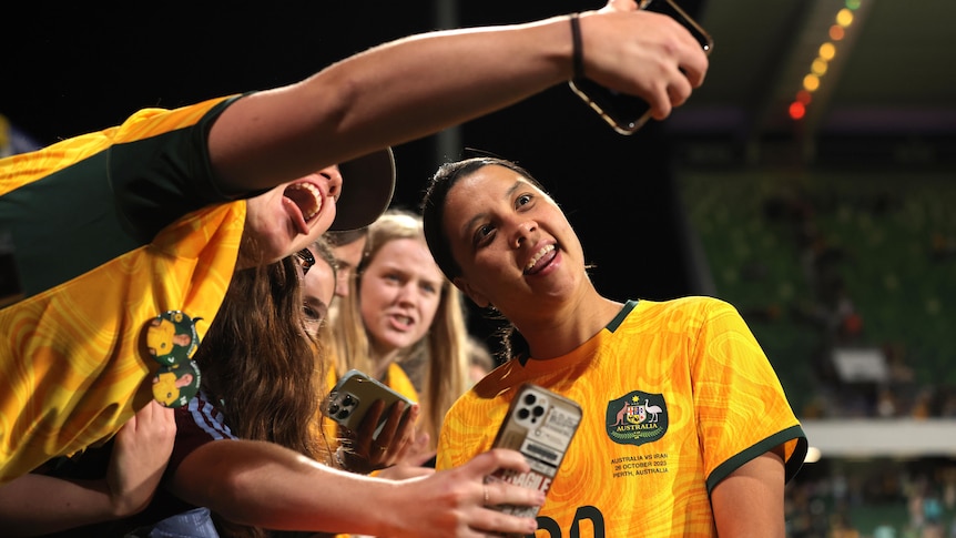 A woman in a yellow soccer shirt poses for a selfie with a young supporter. 