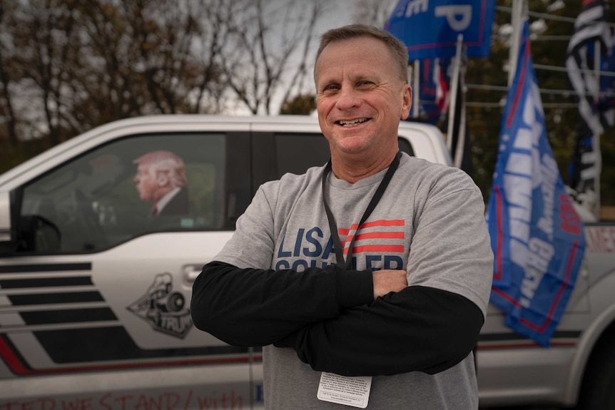 A man smiling with his arms folded across his chest in front of a car with Trump's photo in the window