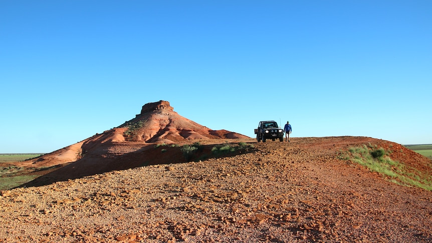 a red earth plain with a red mountain in the background and a car and person in the distance on a sunny day