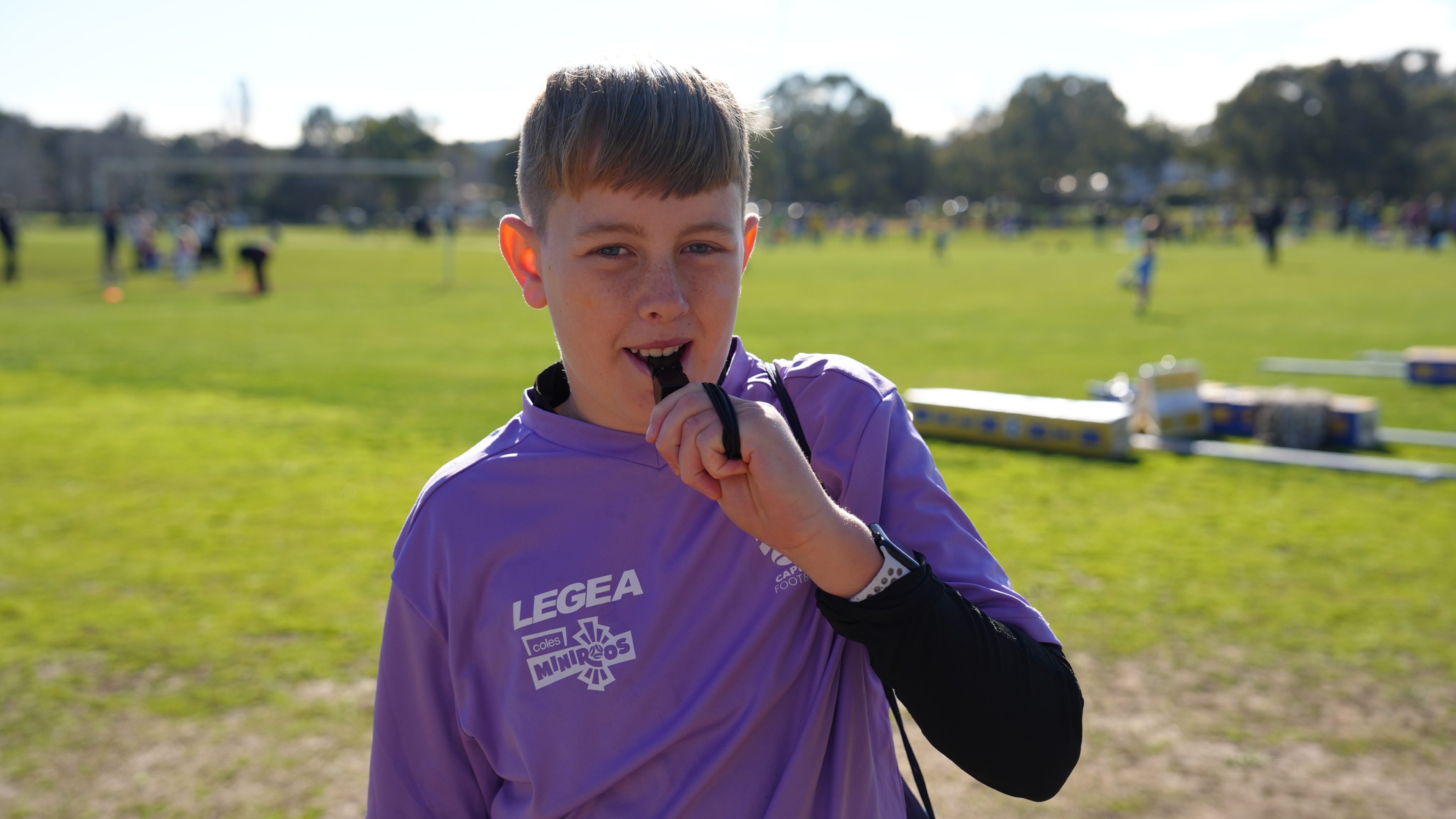 A young boy with short light hair stands on a soccer filed preparing to blow a whistle.