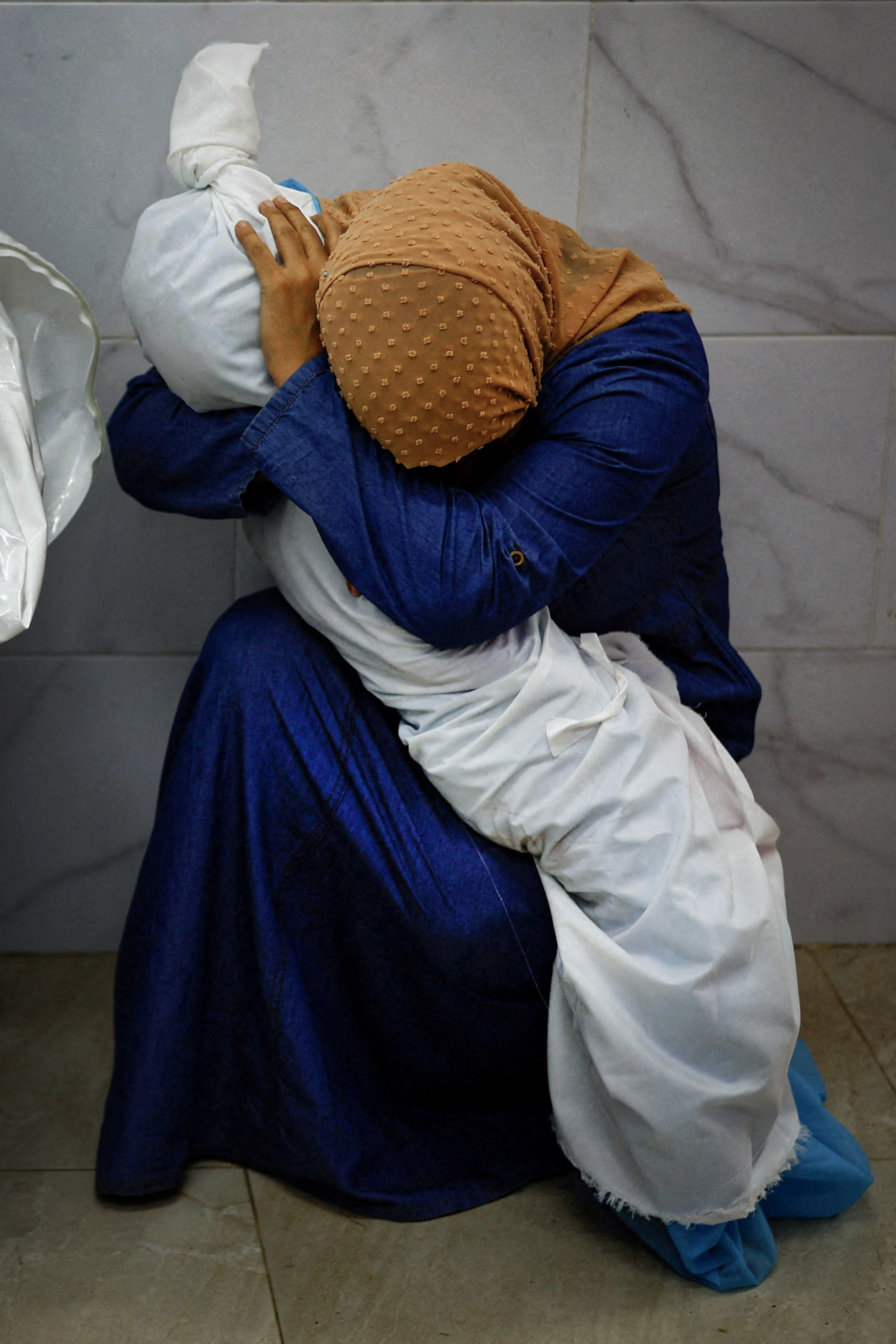 A woman wearing a blue cloak and yellow headscarf crouched on the crowd, holding the body of a small child wrapped in white