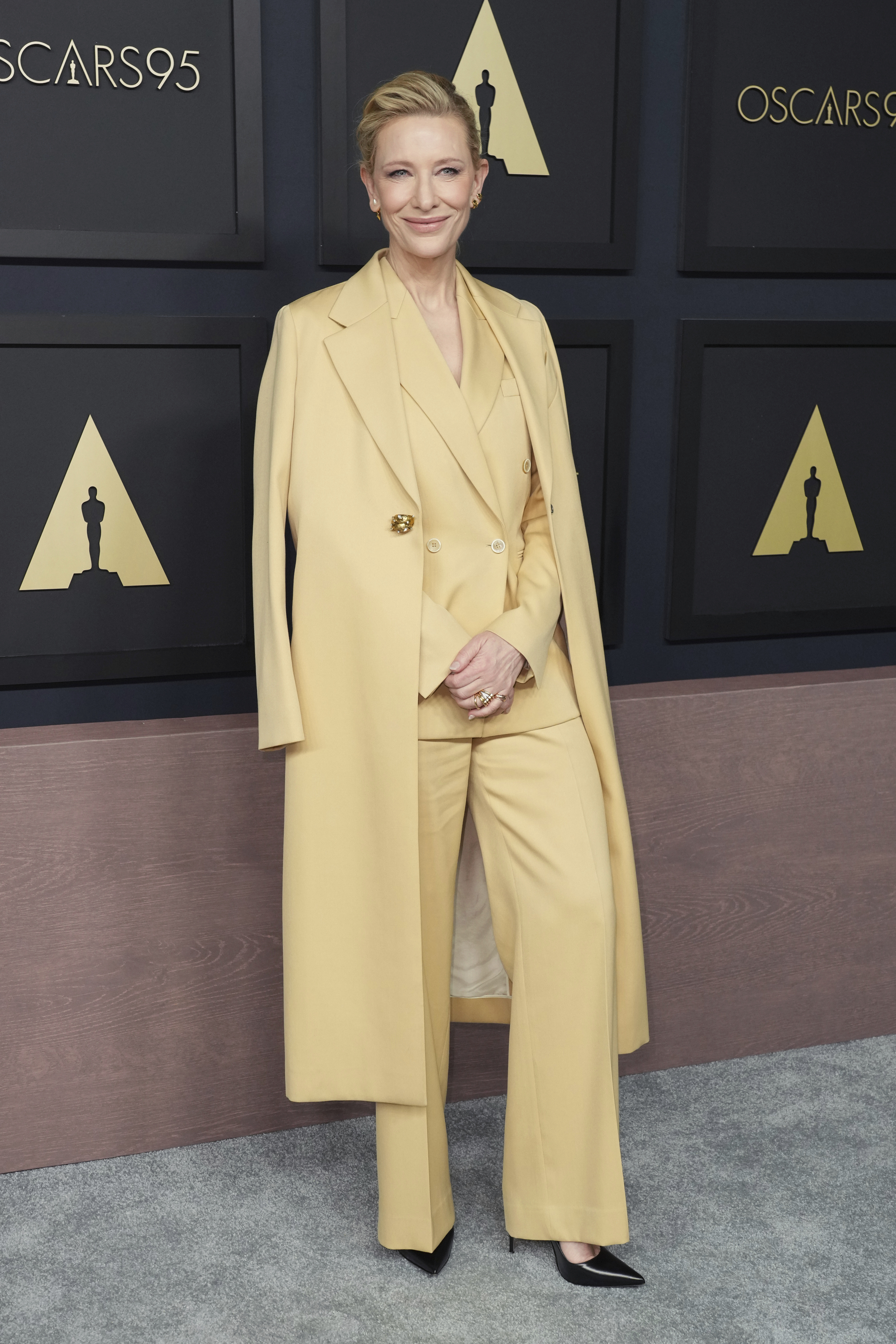 Cate Blanchett wearing a pale yellow pantsuit with a long coat in the same colour draped over her shoulders