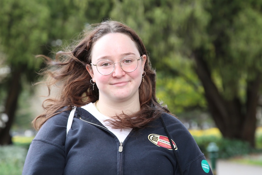 A woman wearing a dark jumper with glasses and long hair, looking into the camera.