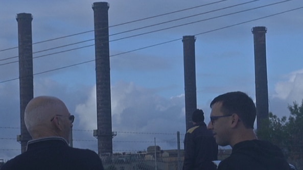 Three men stand behind a wire fence looking out over four tall chimney stacks.