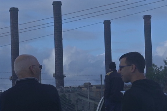 Three men stand behind a wire fence looking out over four tall chimney stacks.