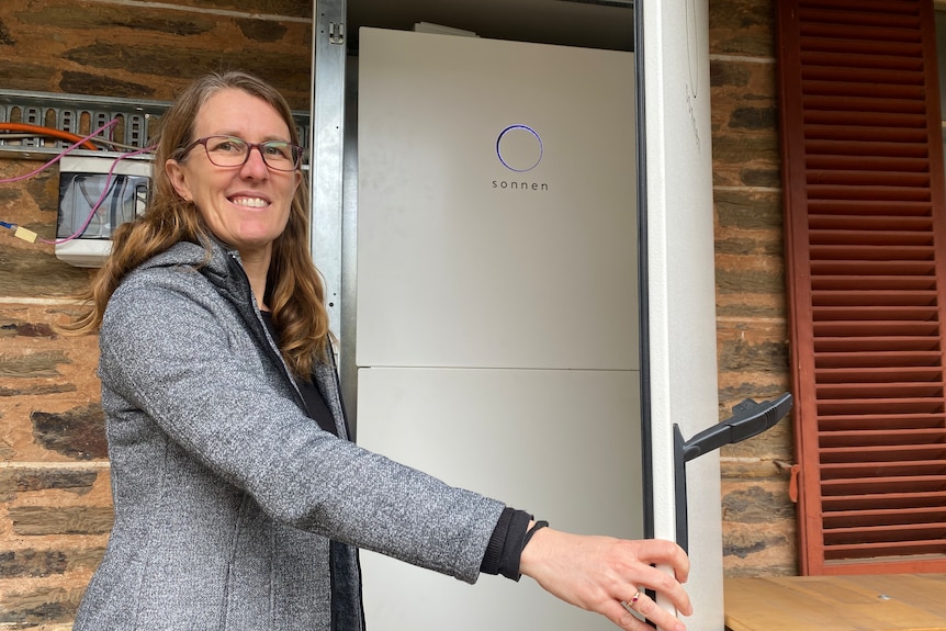 A woman with long brown hair and glasses stands next to a home battery