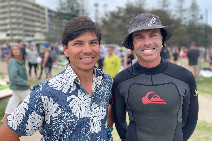 Two men stand on grass, with a crowd behind them. One man is in a floral shirt and one in a wetsuit.