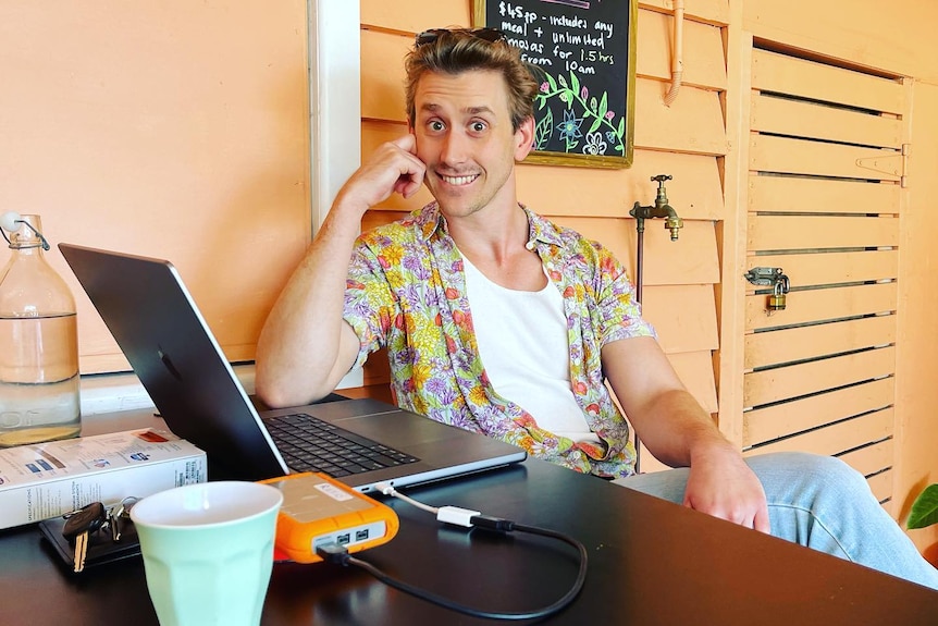 A man wearing a white undershirt, open yellow shirt and jeans sitting at a table in a cafe with a laptop open in front of him