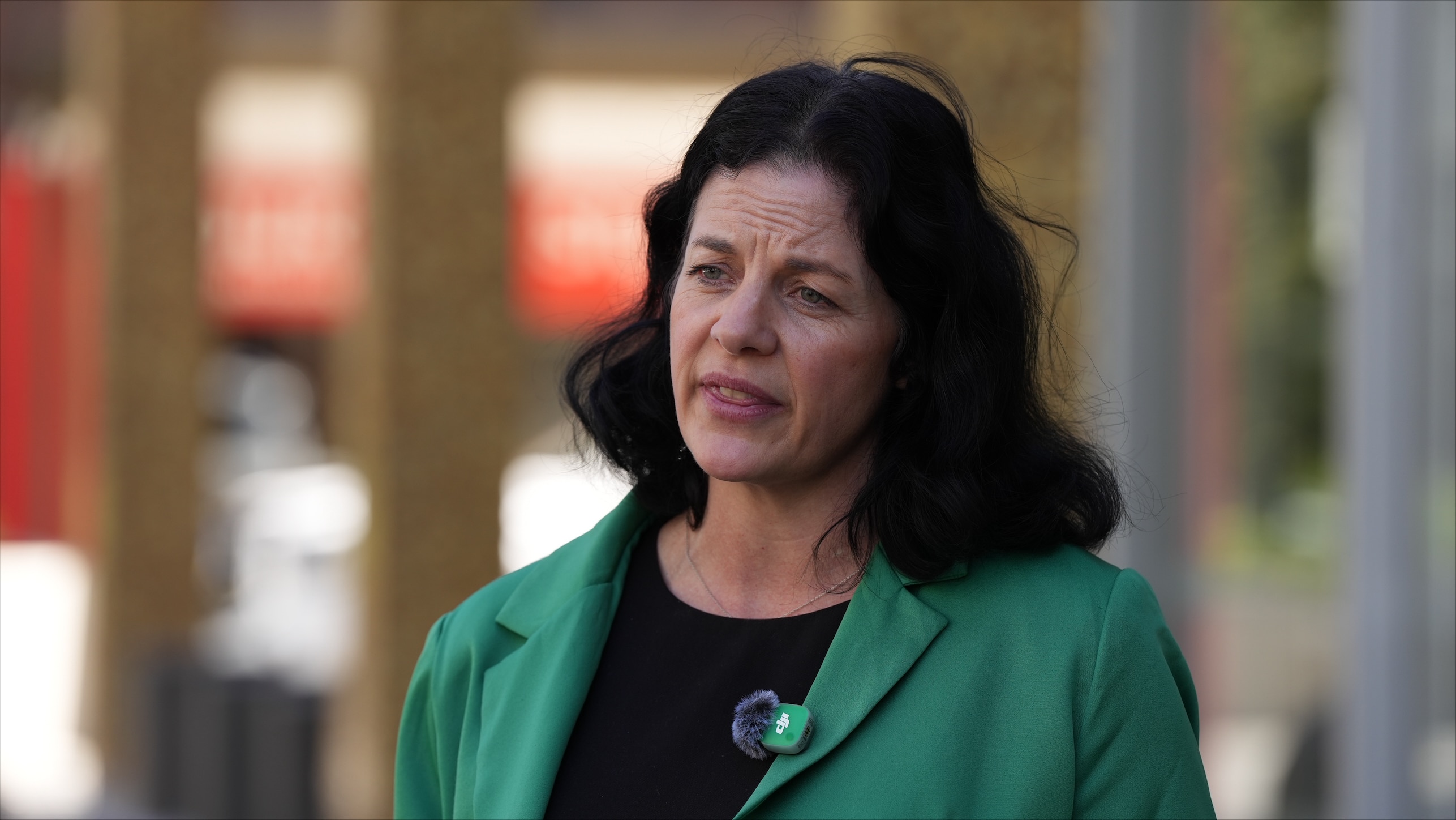 A woman with black shoulder-length wavey hair in a black top and green blazer speaks to media.