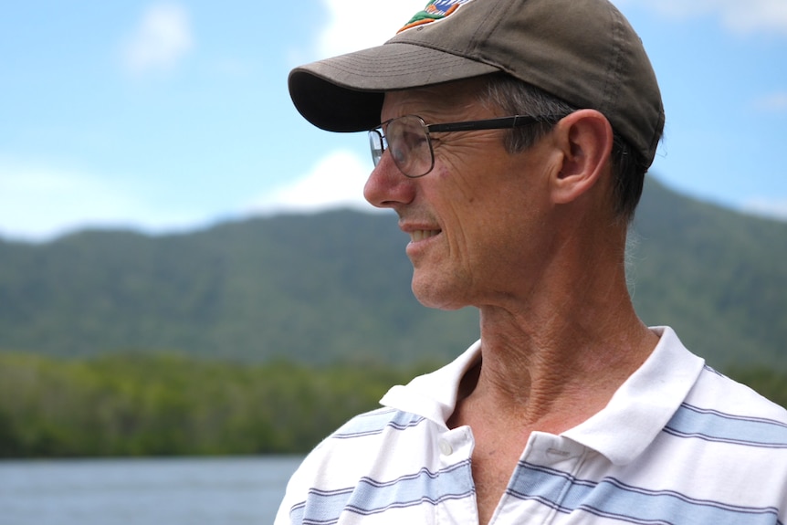 A man, wearing a cap stands next to a river and mountains.