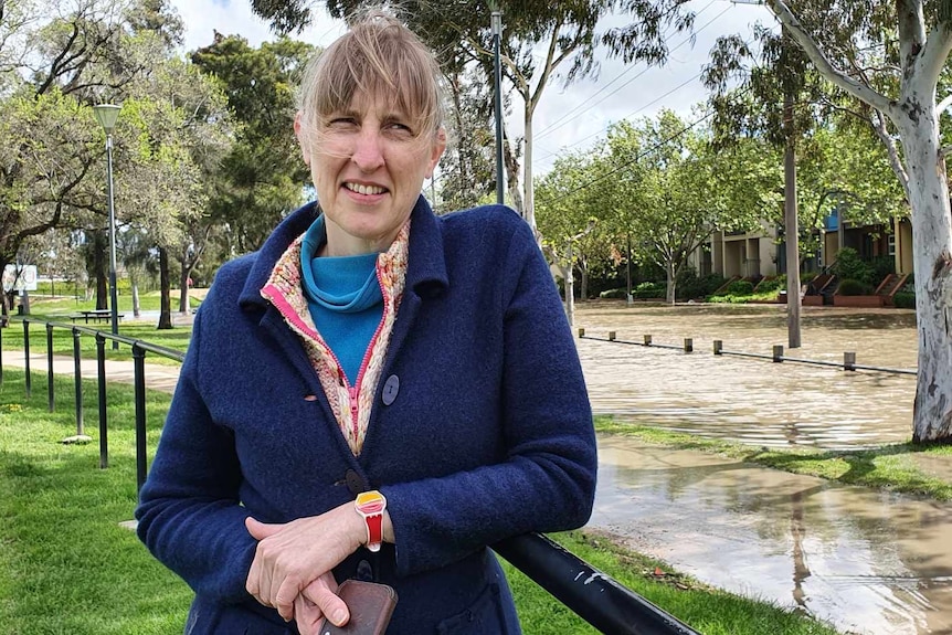 A woman wearing a blue jacket stands in front of a flooded road.