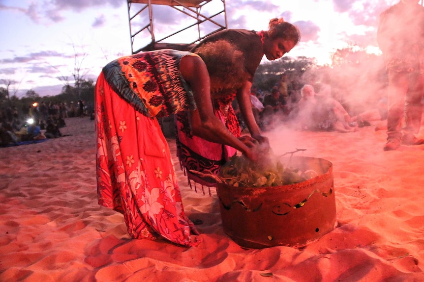 Two aboriginal women lean over a fire drum that is full of leaves that are smoking.