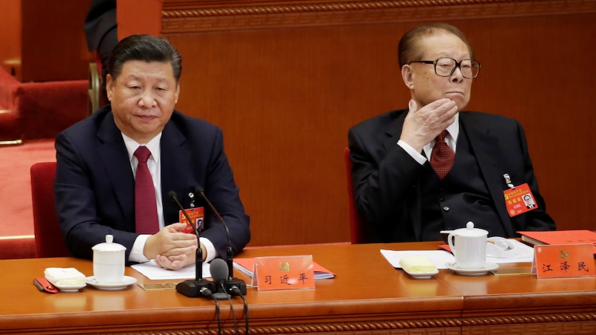 Xi Jinping and Jiang Zemin, both wearing suits with maroon ties, sit at desks. Xi folds his hands while Jiang scratches his chin
