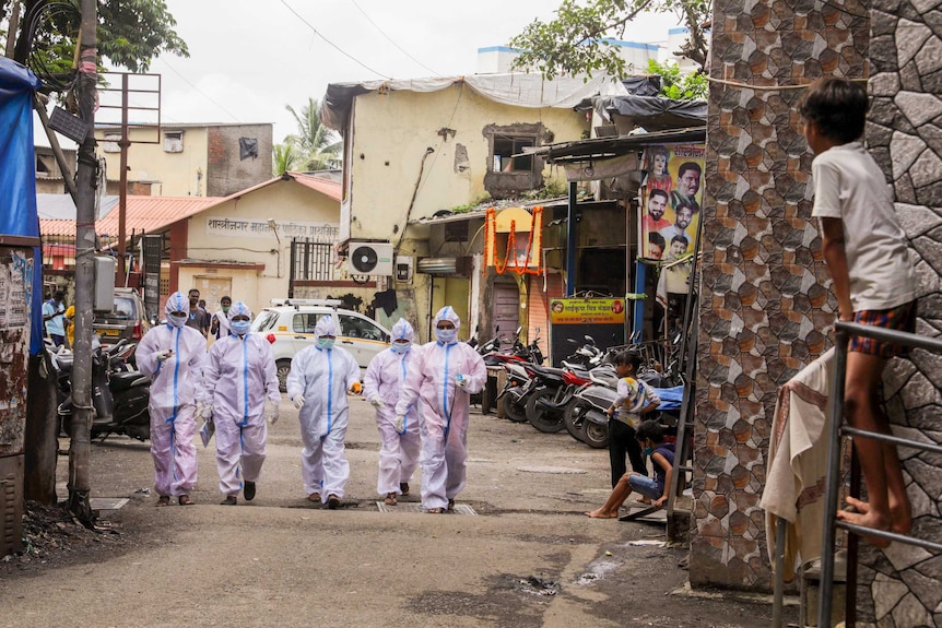 A group of people in full PPE walk through a road in Dharavi while children watch on