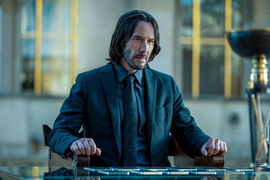 White man with shoulder-length black hair wears a dark teal suit and sits with a quizzical expression at a glass table.