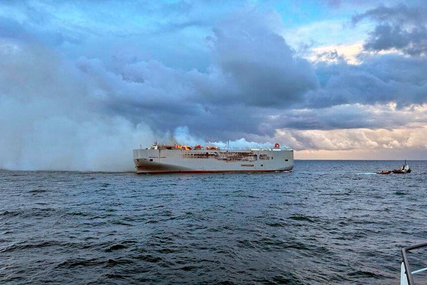 A cargo ship burns in the sea at daytime as smoke rises