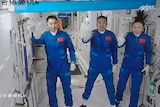 Three people in blue space suits wave from the inside of a space station. They float in zero gravity
