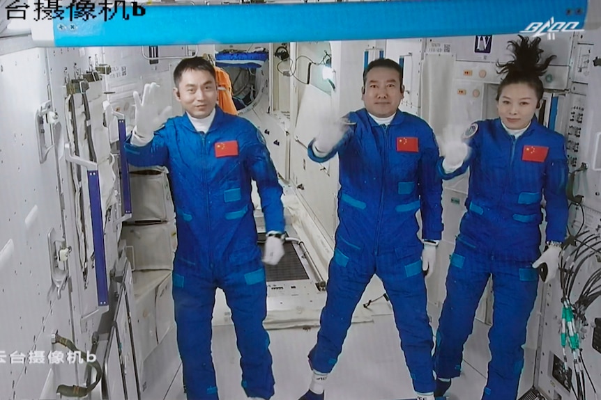 Three people in blue space suits wave from the inside of a space station. They float in zero gravity
