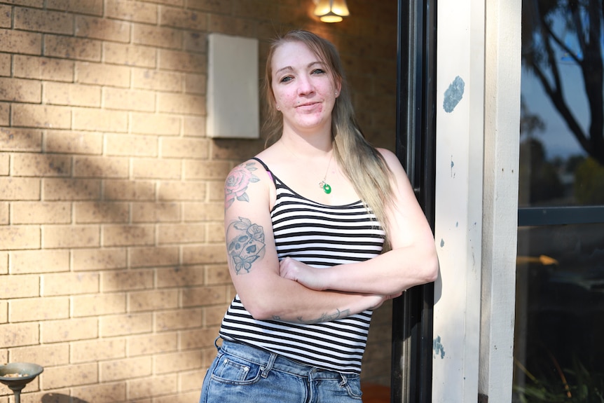 A young woman leans against a rotten wooden beam at the entrance to her home.