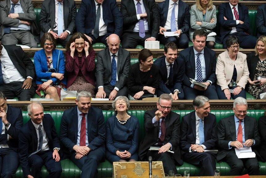 Theresa May laughs surrounded by members of parliament in the House of Commons.