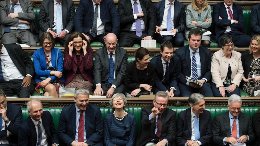 Theresa May laughs surrounded by members of parliament in the House of Commons.