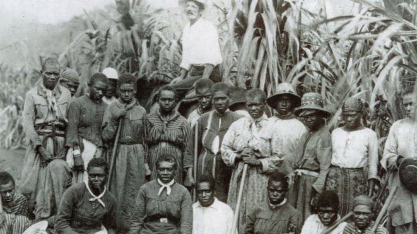 Black and white photo of a mena and women from Vanuatu with a white man standing behind them.