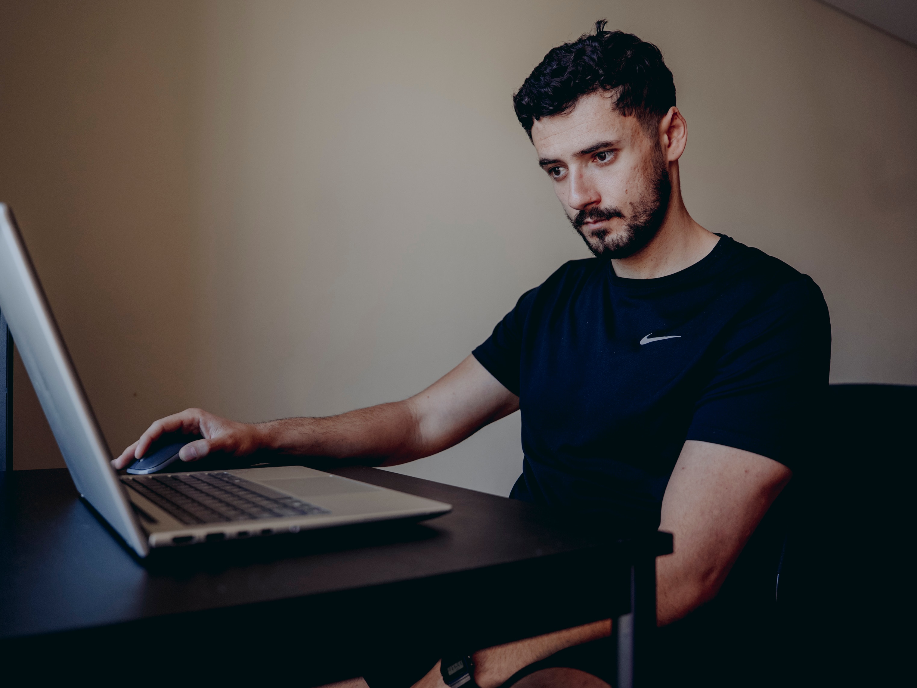 Andre sits at his desk look at his open laptop screen wearing a dark t-shirt. 