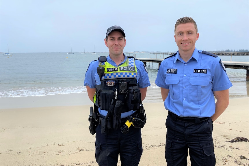 Two police officers in uniform stand on a beach in front of a jetty.