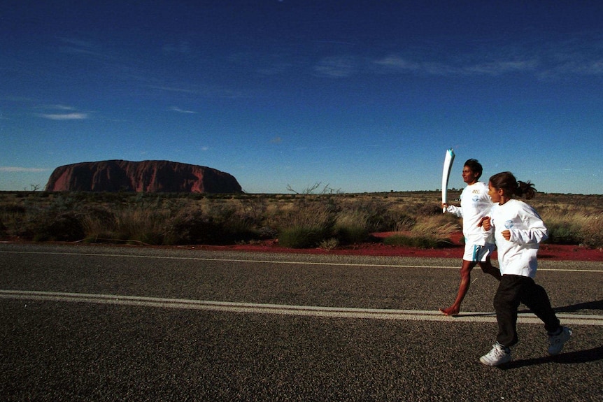 Nova Peris runs barefoot on a road with the Olympic torch. Uluru is in the distance.